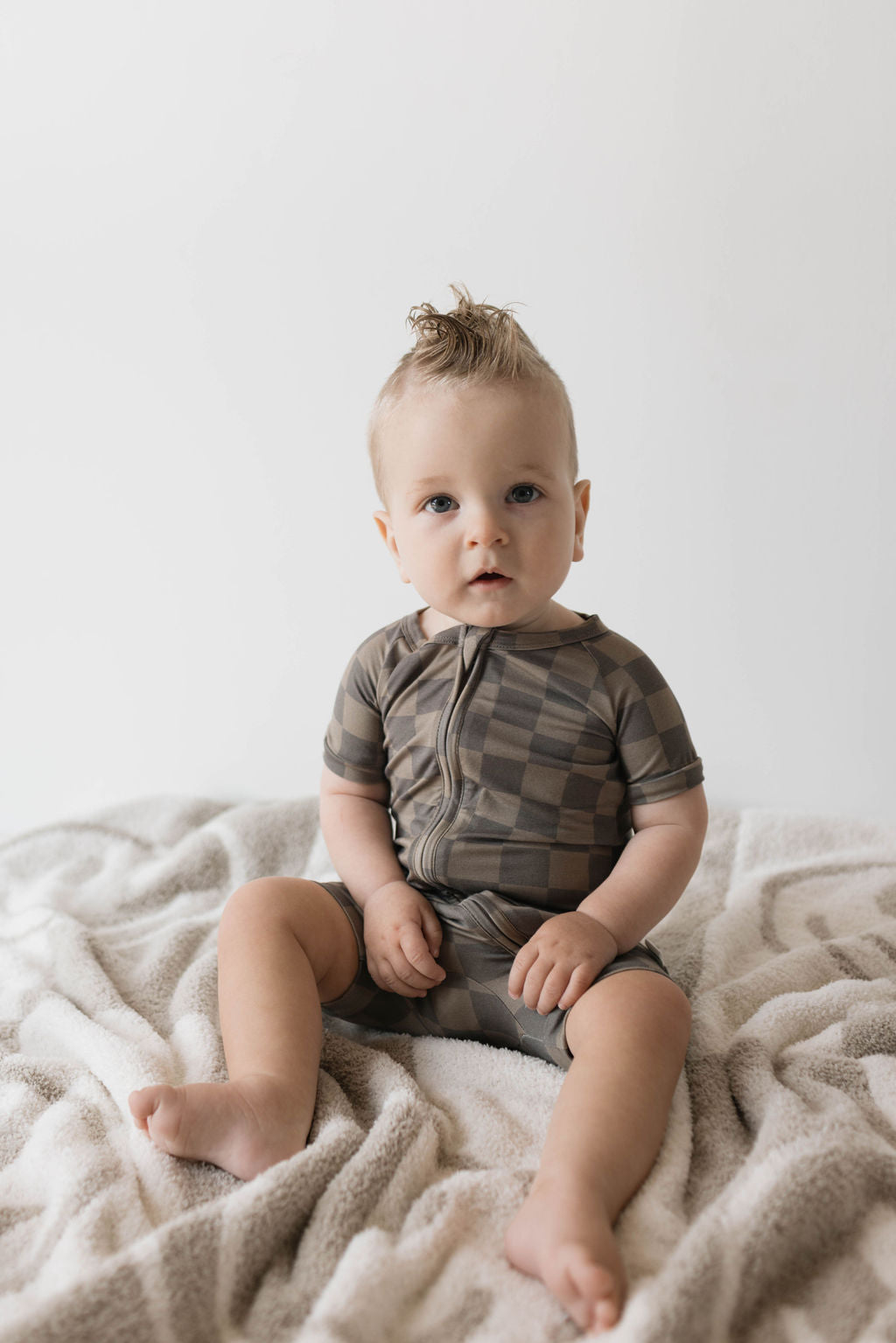 A baby with light hair sits on a soft, beige blanket, wearing the "Shortie Bamboo Zip | Faded Brown Checkerboard" onesie from forever french baby. The breathable bamboo clothing features a checkered pattern and short sleeves. The baby looks toward the camera with a curious expression against a plain, light-colored background.