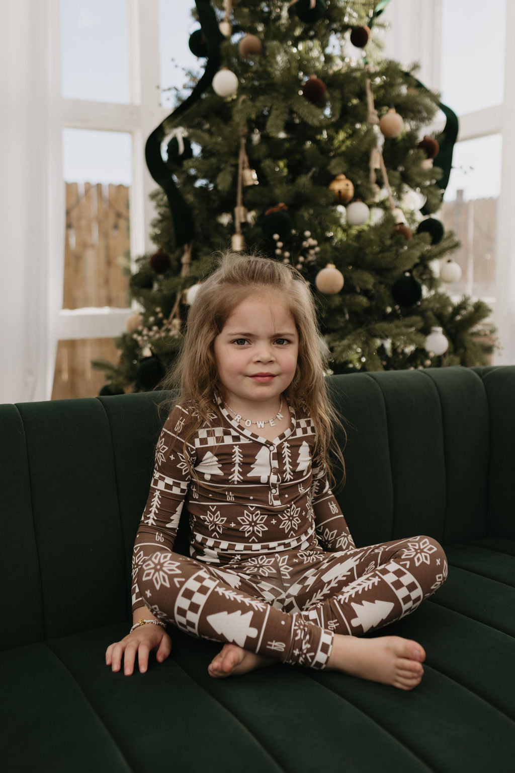 A little girl in forever french baby's breathable Bamboo Two Piece Pajamas | Forever Fair Isle sits cross-legged on a green couch, with a decorated Christmas tree adorned with ornaments in the background, enhancing the festive charm.