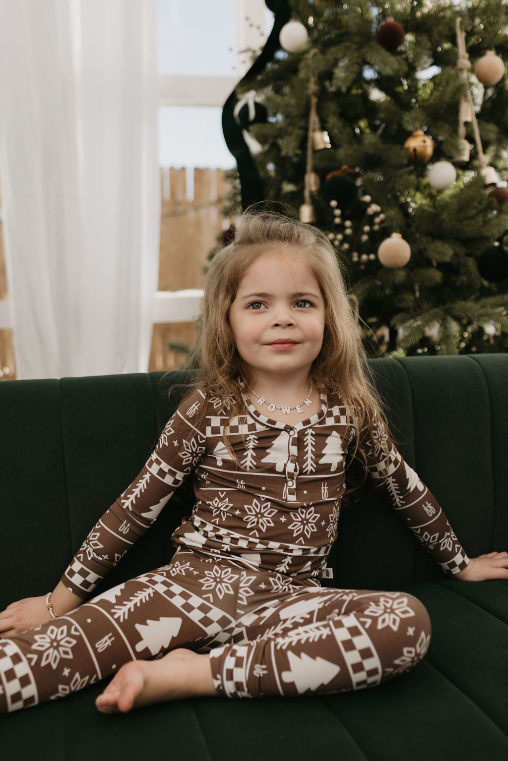 A young child with long hair sits on a dark green couch wearing the breathable "Bamboo Two Piece Pajamas" by forever french baby, featuring festive brown and white designs with tree and snowflake patterns. A decorated Christmas tree is behind them, adding to the cozy holiday atmosphere.