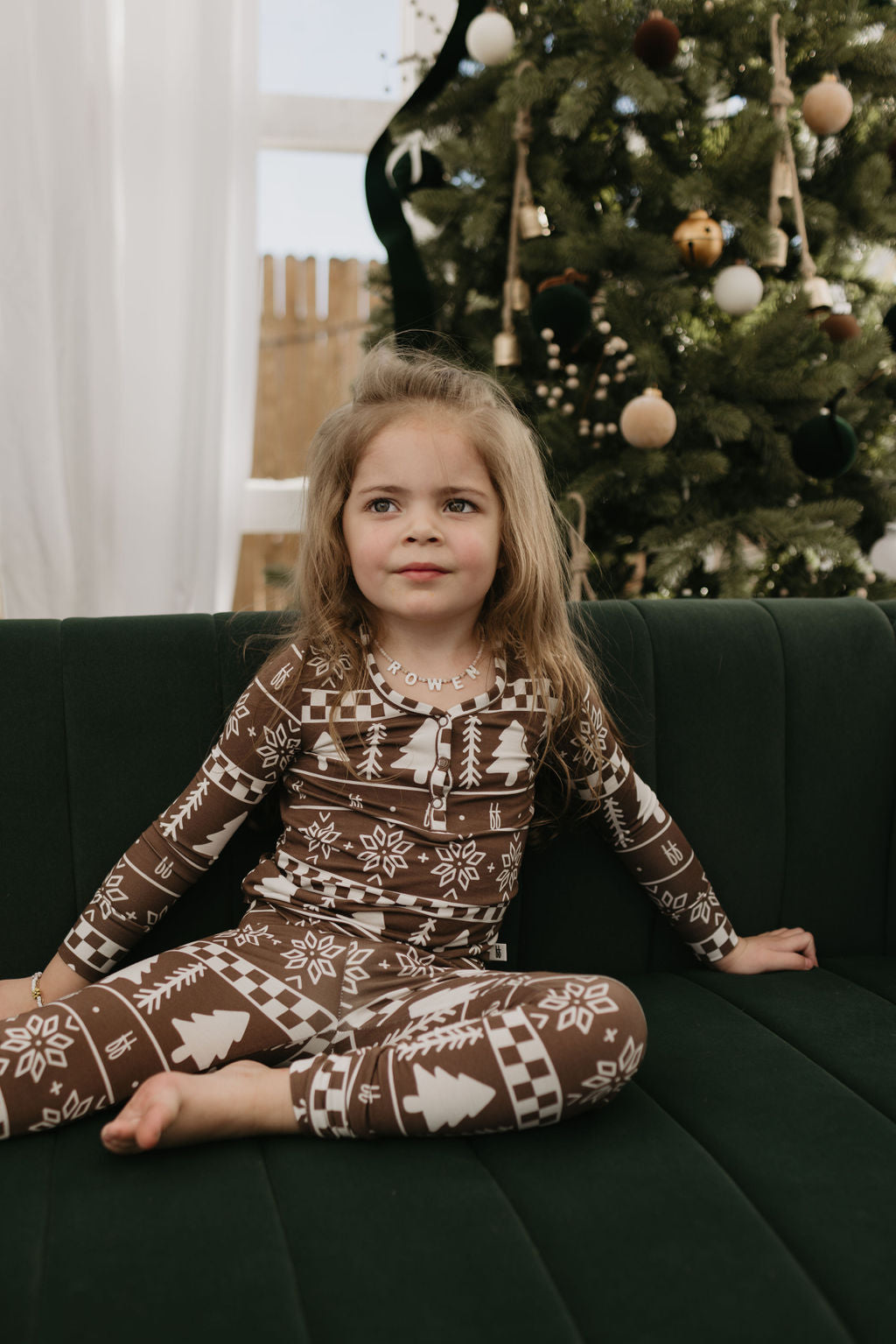 A young girl with long hair, wearing the Forever Fair Isle Bamboo Two Piece Pajamas by forever french baby, sits on a dark green couch. A decorated Christmas tree in the background enhances the cozy ambiance.