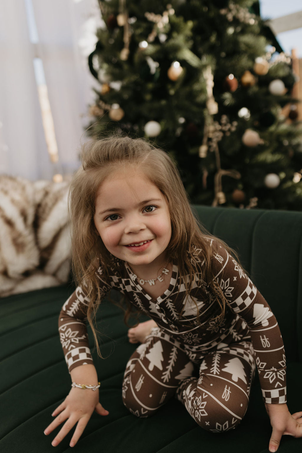 A young child with long brown hair is smiling while sitting on a green couch, dressed in the cozy Bamboo Two Piece Pajamas from forever french baby. The breathable fabric features a charming Forever Fair Isle design in brown and white hues. In the background, a decorated Christmas tree adds to the festive atmosphere.