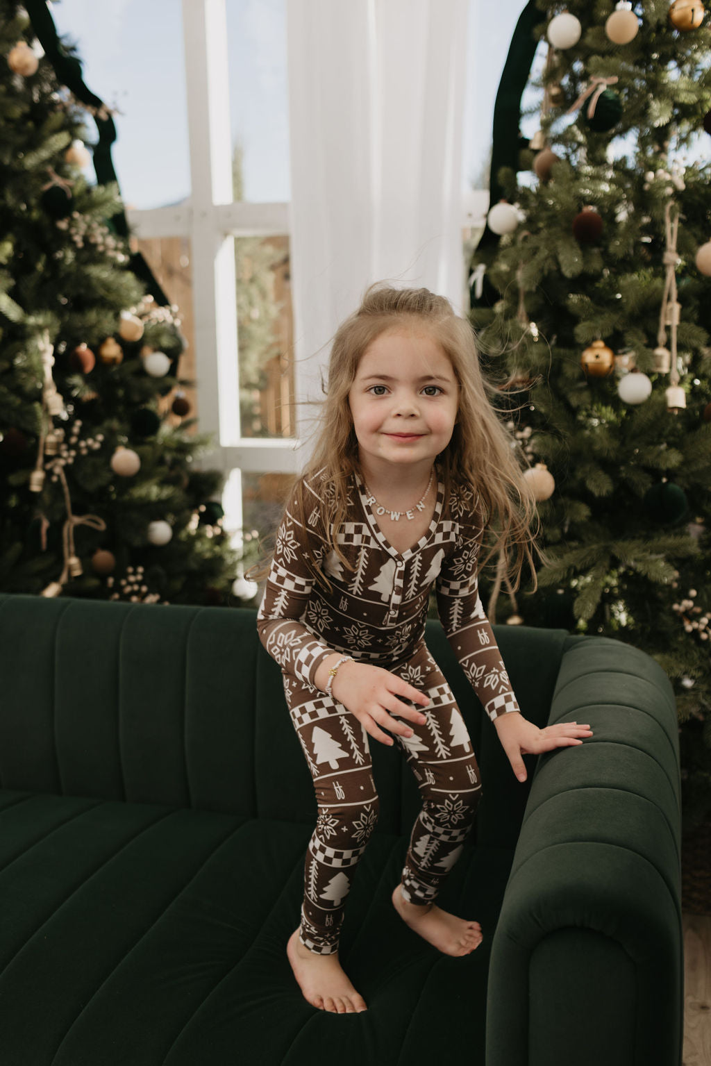 A young girl in the Bamboo Two Piece Pajamas from forever french baby is standing on a dark green sofa, enveloped by beautifully decorated Christmas trees. The festive atmosphere exudes coziness and invitation, while her breathable Forever Fair Isle outfit ensures she remains comfortable amid the holiday cheer.