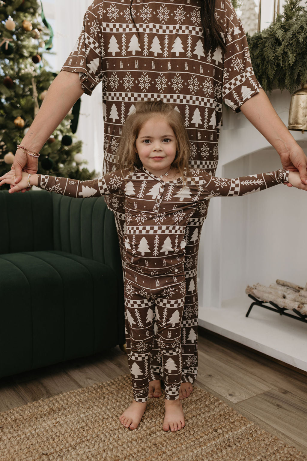 A child and an adult are wearing matching Forever Fair Isle bamboo pajamas by forever french baby, featuring festive white Christmas-themed patterns. The child joyfully extends their arms while being held by the adult. In the background, a Christmas tree and fireplace create a cozy holiday setting.