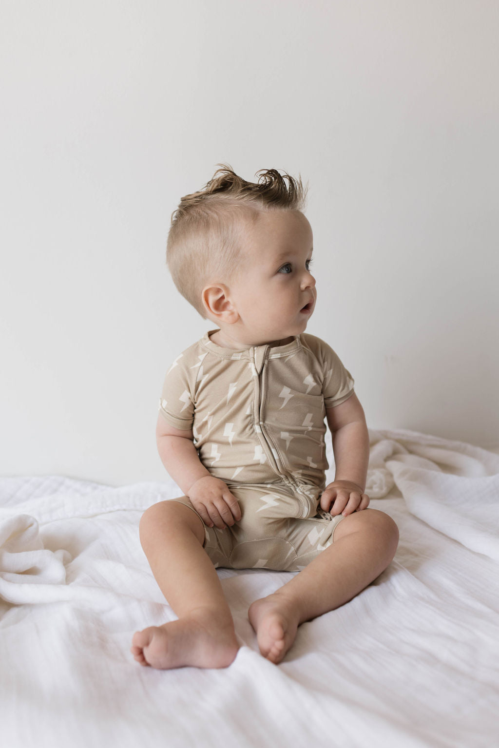 A baby with light hair sits on a white blanket against a plain background, wearing the "Shortie Bamboo Zip | Tan Lightning Bolt" from Forever French Baby. The hypo-allergenic pajamas feature a beige color with small white lightning bolt patterns that ensure a cozy fit as the baby gazes off to the side with a curious expression.