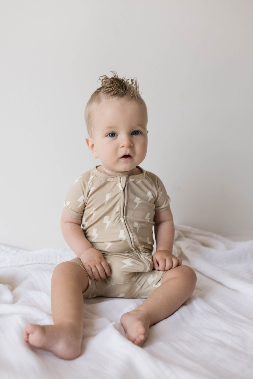 A baby with a light-colored mohawk hairstyle sits on a white blanket against a plain background. The baby is wearing the "Shortie Bamboo Zip | Tan Lightning Bolt" romper from forever french baby, which is hypo-allergenic and features white lightning bolt patterns. The baby looks towards the camera with a neutral expression.