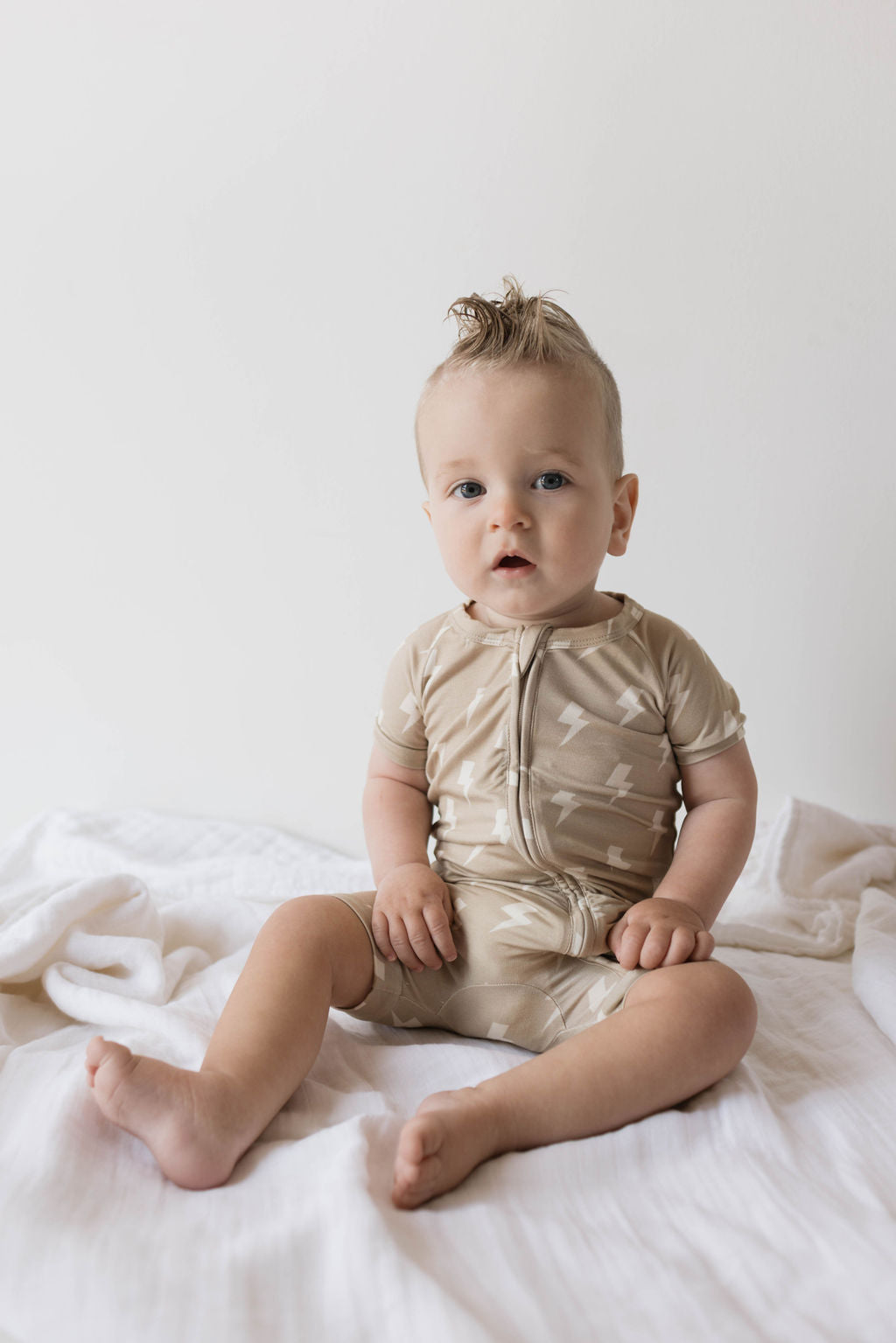 A baby with light hair sits on a white blanket against a plain background, wearing the forever french baby's Shortie Bamboo Zip in Tan Lightning Bolt pajamas. The baby has a curious expression and looks directly at the camera.