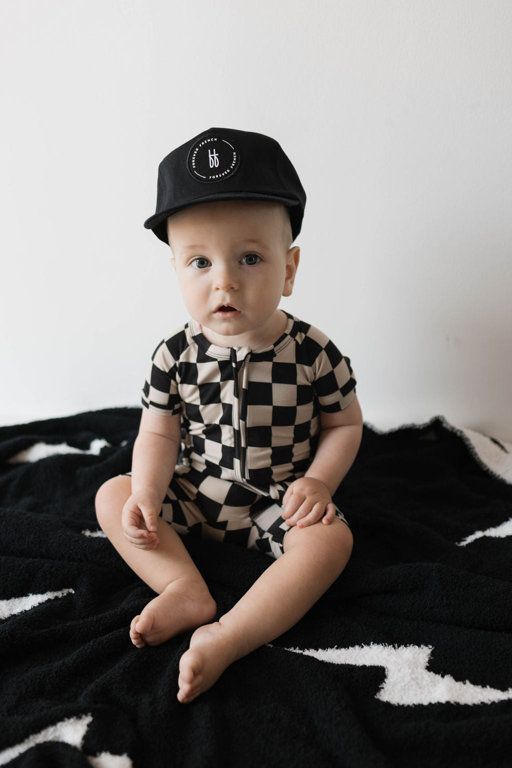 A baby sits on a black and white blanket, dressed in hypo-allergenic Shortie Bamboo Zip clothing in a black checkerboard pattern from forever french baby, accessorized with a black hat featuring a circular logo. The baby looks at the camera with a neutral expression.