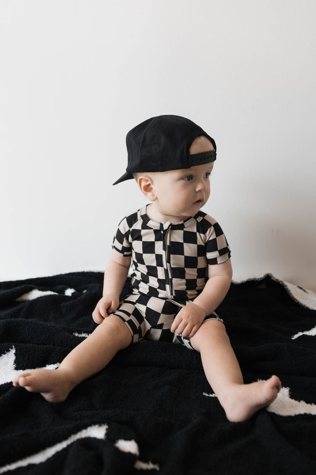 A baby sits on a black blanket adorned with white star patterns, dressed adorably in the hypoallergenic forever french baby Shortie Bamboo Zip in Black Checkerboard and a backward black cap. The background is a simple white wall, and the baby gazes to the side with a serene expression.