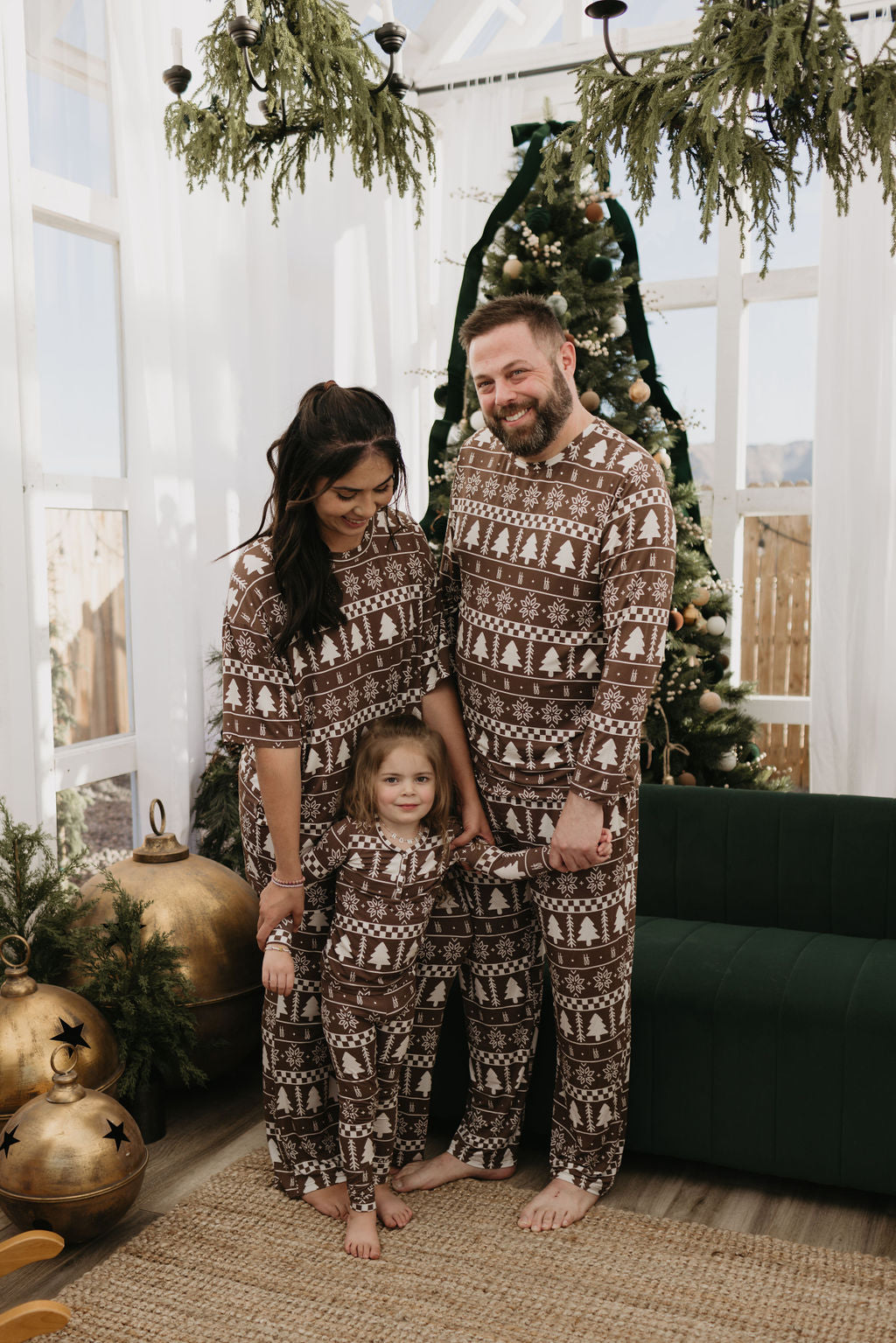 A family of three, all dressed in Forever French Baby's Short Sleeve Women's Bamboo Pajamas in the Forever Fair Isle design, stands together in a decorated living room featuring a Christmas tree and large ornaments. The room is filled with bright natural light.