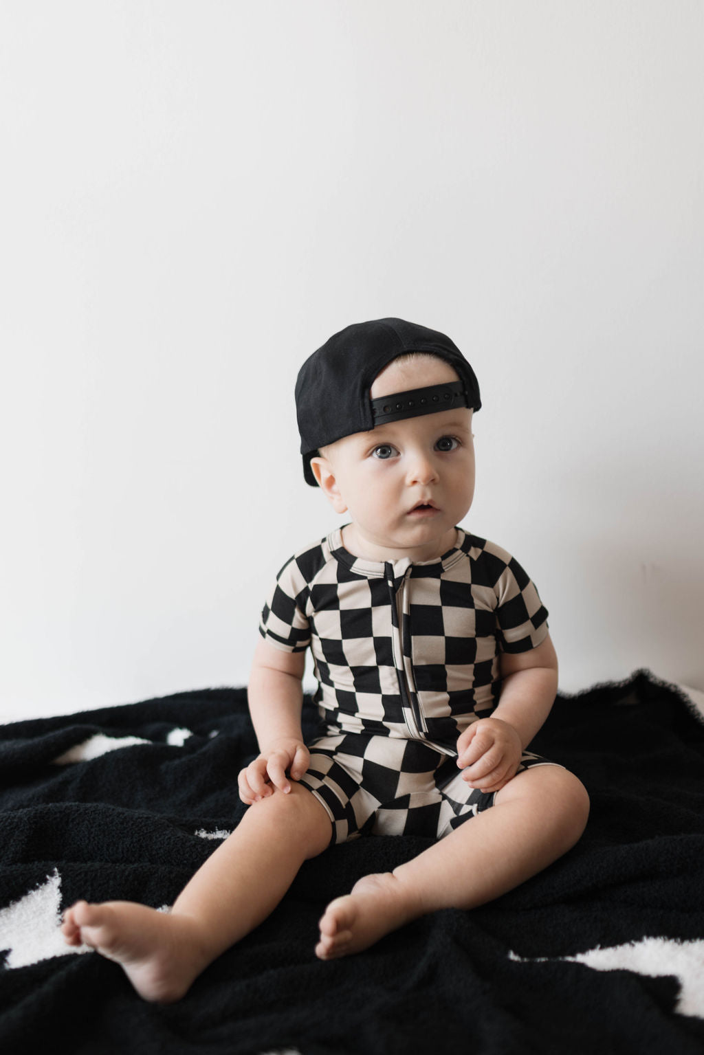 A baby wearing the forever french baby's Shortie Bamboo Zip in Black Checkerboard, made from hypo-allergenic fabric, and a black cap sits on a black blanket with a white background. The baby is looking slightly to the side with a curious expression.