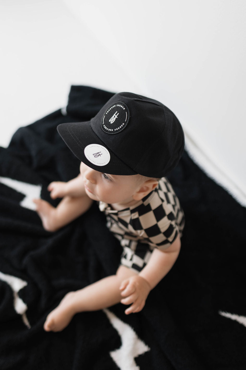 A baby from the forever french baby collection sits on a black and white blanket, wearing the Children’s Trucker Hat | ff Black with a round patch and matching checkered outfit. The baby appears curious, looking out of the frame. Care instructions recommend spot cleaning only.