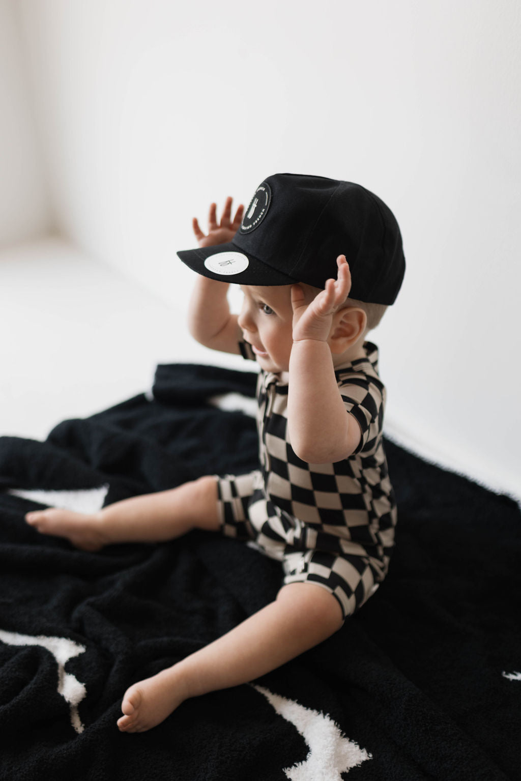 A baby, clad in the "Shortie Bamboo Zip | Black Checkerboard" from forever french baby, sits on a black blanket adorned with white star patterns. The baby is wearing a black cap and has its hands raised towards the cap, seemingly adjusting it. The backdrop is simple and white.