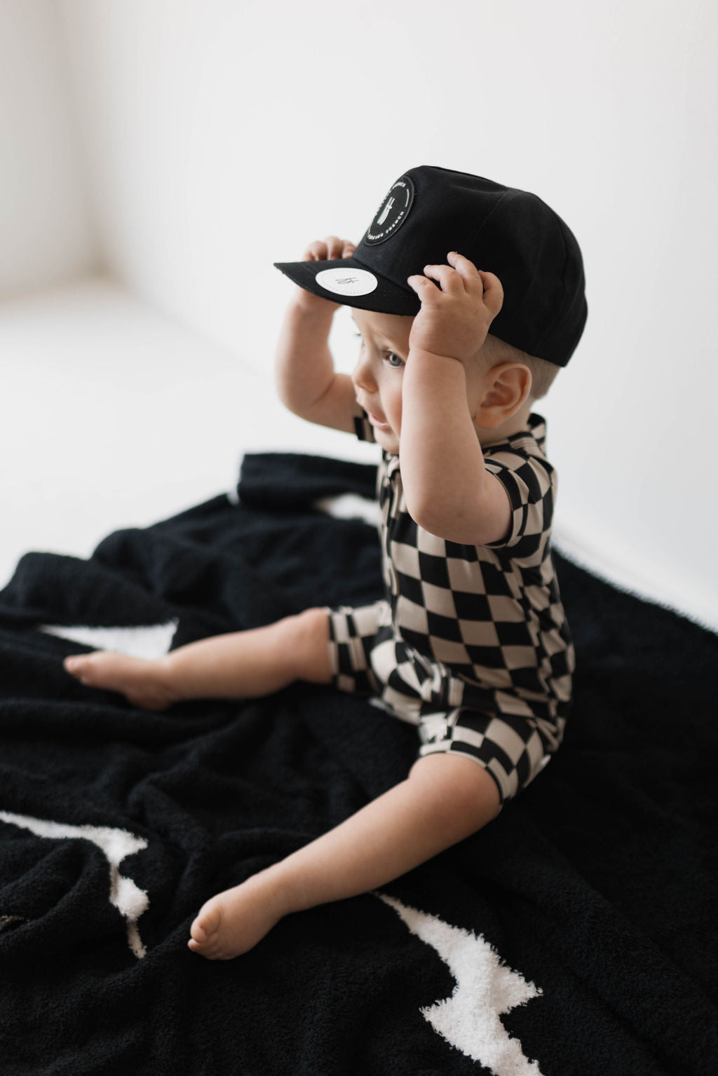 A baby sits on a black and white blanket, clad in the forever french baby Shortie Bamboo Zip in Black Checkerboard, accessorized with a black cap. The baby is holding the brim of the cap with both hands and looking upward against a minimal, light-colored background.