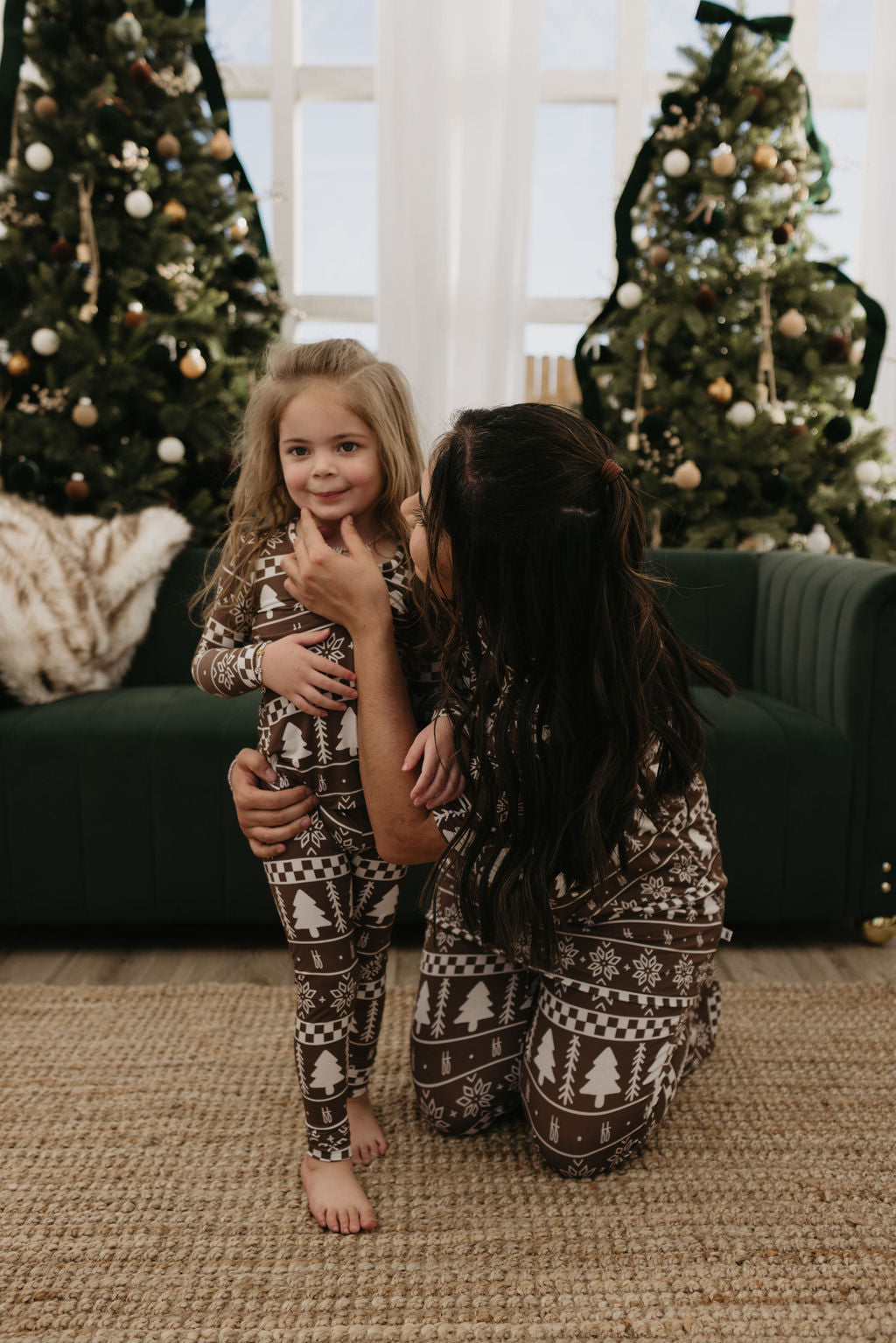 A woman and a young girl are joyfully wearing matching Forever Fair Isle bamboo two-piece pajamas from forever french baby, standing before a festive couch surrounded by decorated Christmas trees. The breathable fabric ensures comfort as the woman playfully holds the girl's face, creating an enchanting holiday scene.