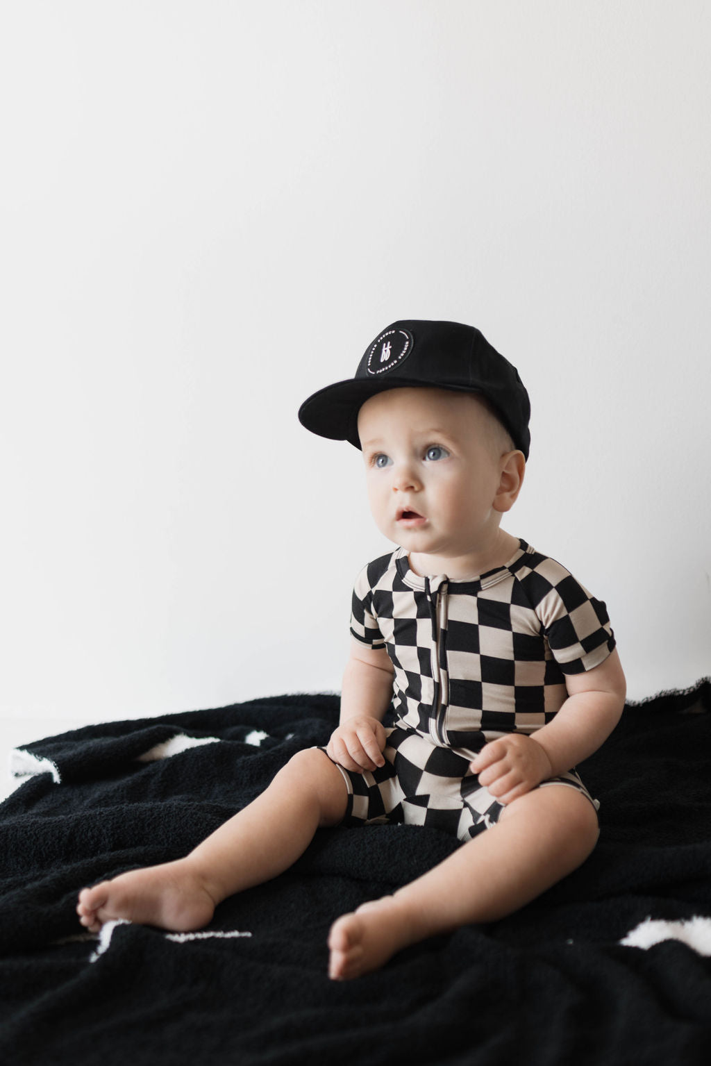 A baby wearing the hypo-allergenic Shortie Bamboo Zip in the Black Checkerboard design from forever french baby and a black baseball cap sits on a soft black blanket adorned with white star patterns, set against a plain white background. The baby looks upward with a curious expression.