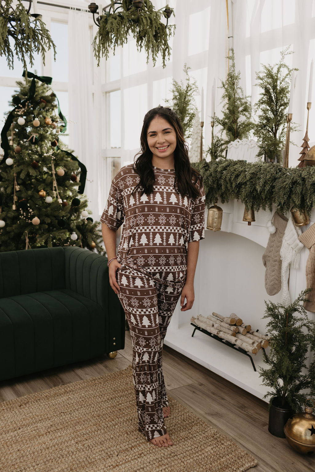 In a cozy living room, a person stands wearing comfortable Short Sleeve Women's Bamboo Pajamas from forever french baby, featuring the Forever Fair Isle design with snowflake and Christmas tree patterns. A decorated Christmas tree glows softly in the background next to a green sofa, while greenery and candles adorn the mantel, creating a warm holiday ambiance.