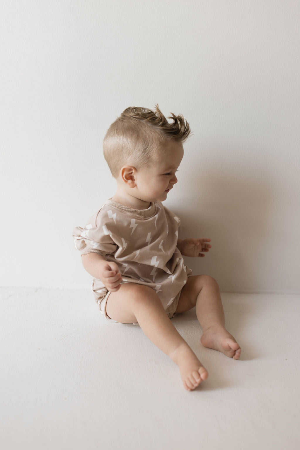 A baby with light-colored hair sits on the floor against a plain, light-colored wall. The baby is wearing a Cream Lightning Sweatshirt Romper by forever french baby and is looking to the side. With cute, styled hair and a relaxed demeanor, this scene captures the essence of muted tones by forever french baby.