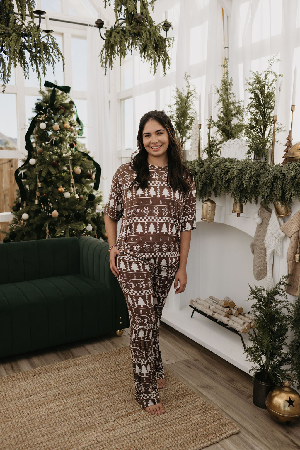 Wearing a Short Sleeve Women's Bamboo Pajama set in Forever Fair Isle by forever french baby, a person stands comfortably in a cozy living room filled with holiday decor, featuring a Christmas tree and garland on the fireplace. A green couch is positioned on the left, with stockings hanging from the mantel.