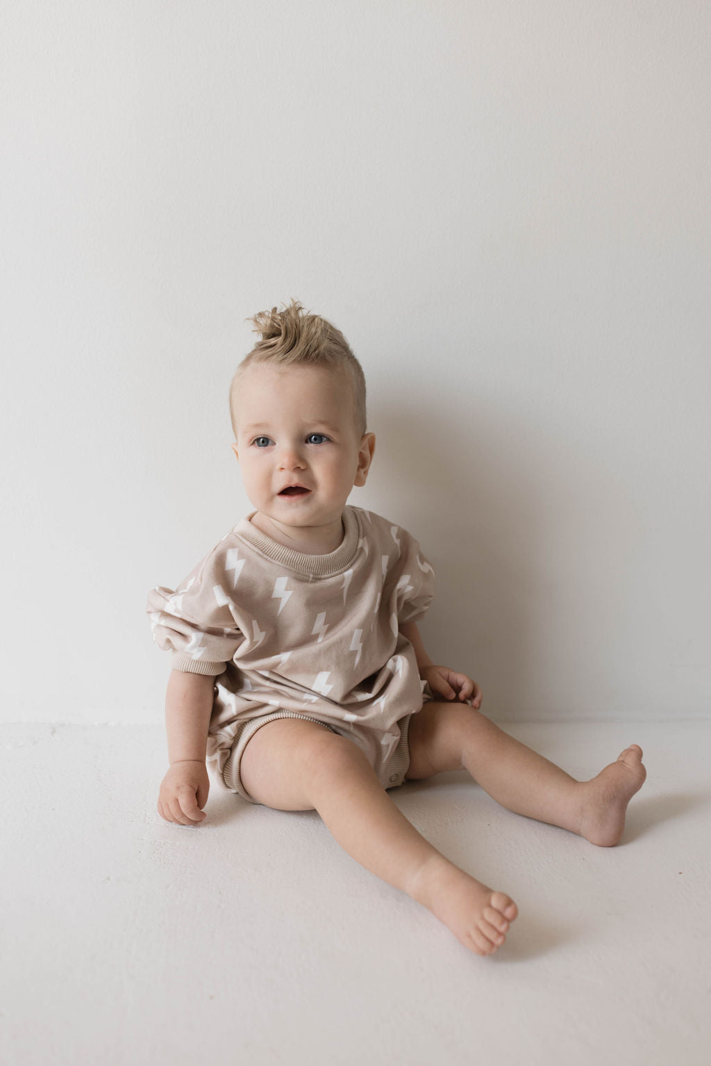 A baby with short, spiky hair sits against a plain beige background. Dressed in a light beige "Sweatshirt Romper | Cream Lightning" by Forever French Baby, the child looks slightly to the side with a small smile. In muted tones, the baby has a relaxed posture with legs stretched out.