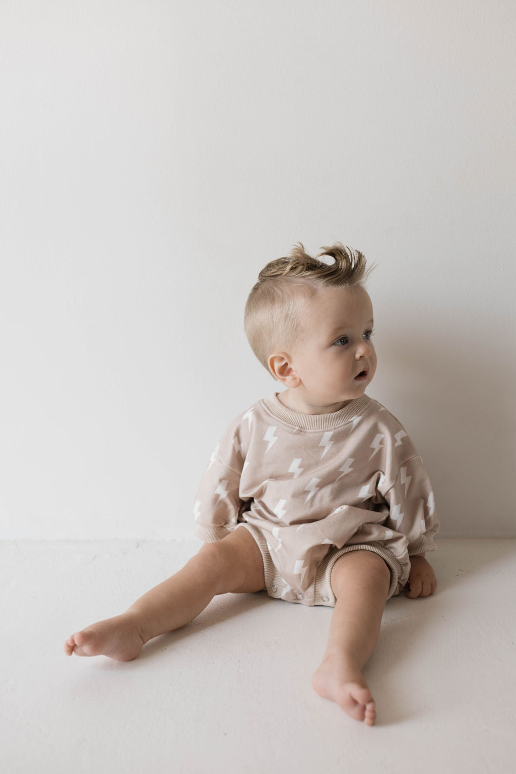 A toddler with short blonde hair sits on the floor against a white wall, donning a light beige Sweatshirt Romper | Cream Lightning adorned with white lightning bolt patterns. The child, dressed in Forever French Baby style, looks to the side with an inquisitive expression, legs stretched out in front.