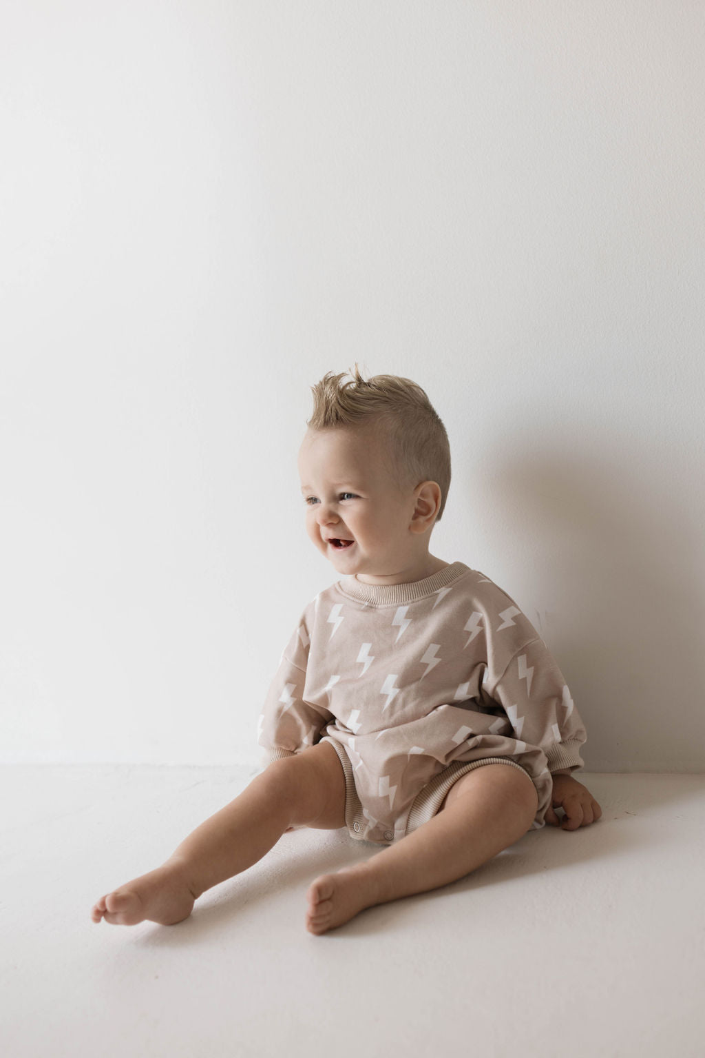 A baby with short, light hair sits on the floor against a white wall. The baby is smiling and wearing a beige "Sweatshirt Romper | Cream Lightning" with white lightning bolt patterns. Clad in these muted tones from forever french baby, the baby appears happy and relaxed.