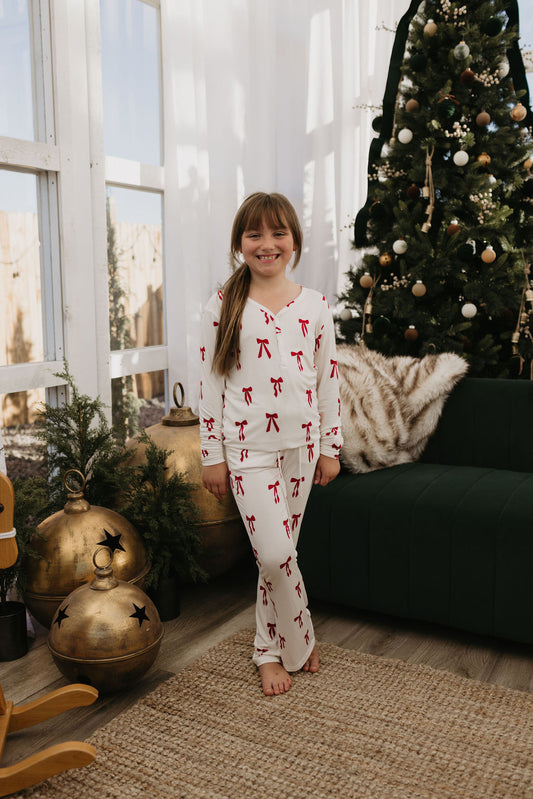 A girl dressed in forever french baby's Pre-Teen Flare Bamboo Pajamas | Ribbons & Bows smiles warmly in a cozy room adorned for Christmas, complete with a Christmas tree, large golden ornaments, and a fur throw draped over a dark green couch, as sunlight pours through the window.