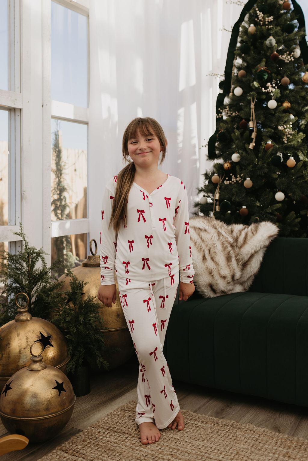 A young girl dressed in "Pre-Teen Flare Bamboo Pajamas | Ribbons & Bows" by forever french baby stands beside a decorated Christmas tree. She is smiling and positioned near large gold ornaments.