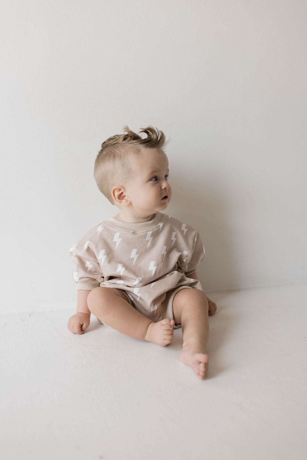 A toddler with light hair and a short haircut sits on the floor, looking to the side. Dressed in the Cream Lightning Sweatshirt Romper from forever french baby, the child embodies the muted tones of Forever French Baby. The background is plain and light-colored.