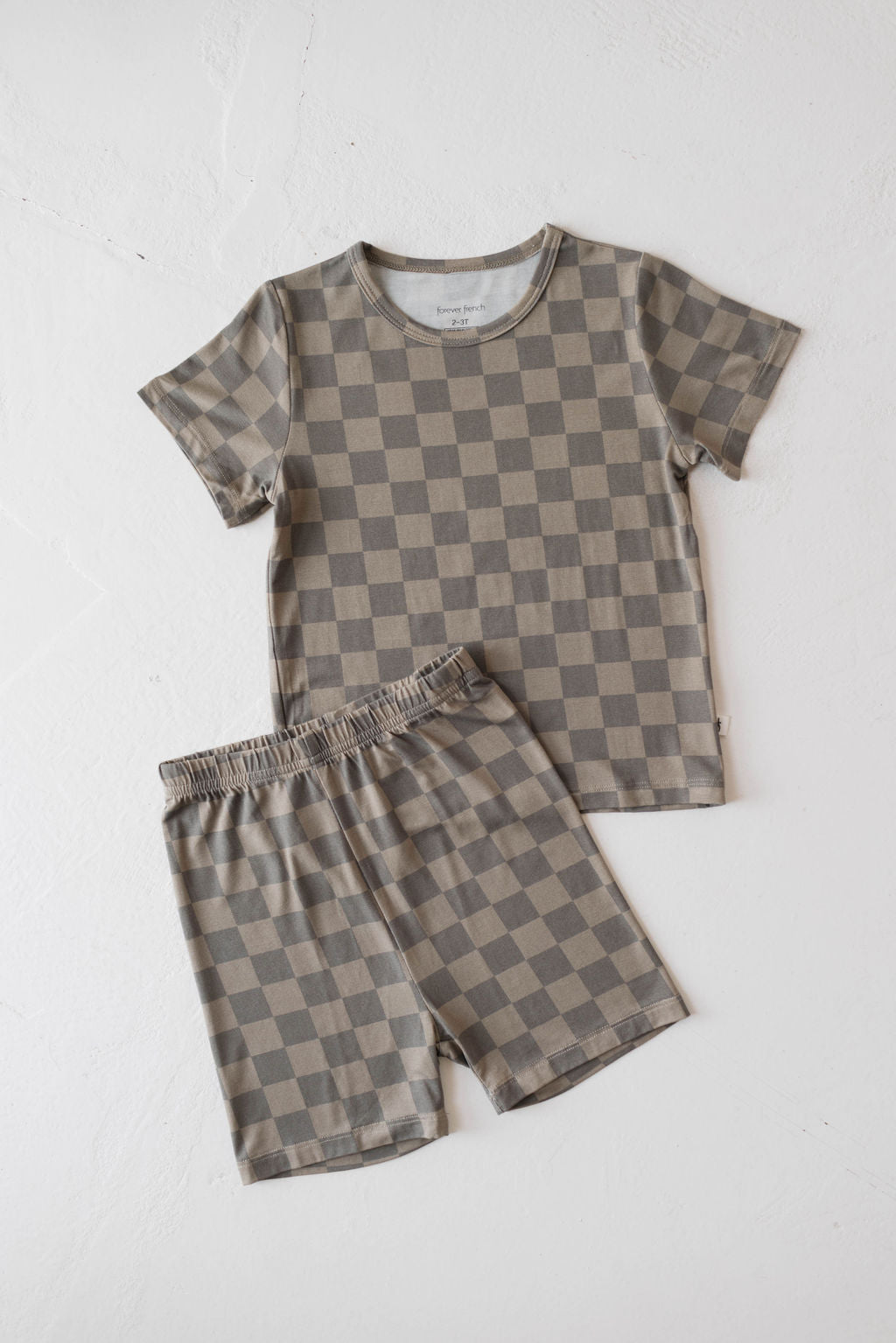 The Bamboo Short Set in Faded Brown Checker by forever french baby is displayed on a white background. This kids' clothing set includes a short-sleeve shirt and matching shorts, both adorned with a brown and beige checkerboard pattern. Made from breathable material, the shorts feature an elastic waistband for added comfort and style.