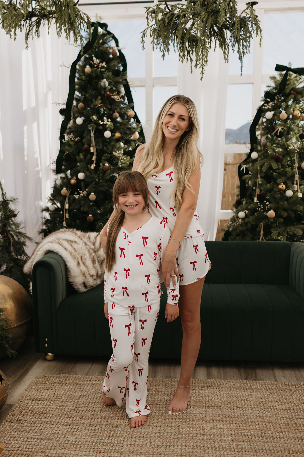 A woman and her pre-teen joyfully pose in matching "Pre-Teen Flare Bamboo Pajamas | Ribbons & Bows" by forever french baby, set in a holiday-adorned room featuring two Christmas trees and lush greenery. A nearby green couch, topped with a cozy blanket, adds to the festive coziness of the scene.