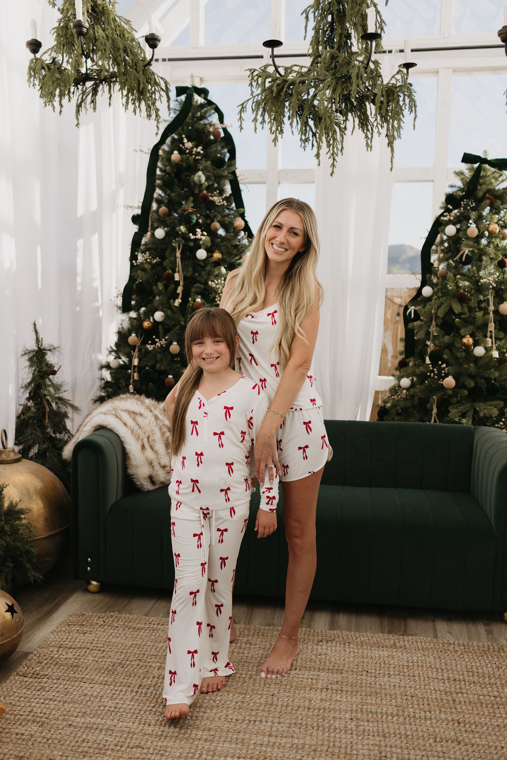 A woman and a young girl stand smiling in a festive setting with Christmas trees behind them. Both are wearing matching Cami Women's Bamboo Sets from forever french baby, crafted from breathable bamboo fabric, adorned with red ribbons and bows. They stand next to a dark green couch surrounded by Christmas decorations.