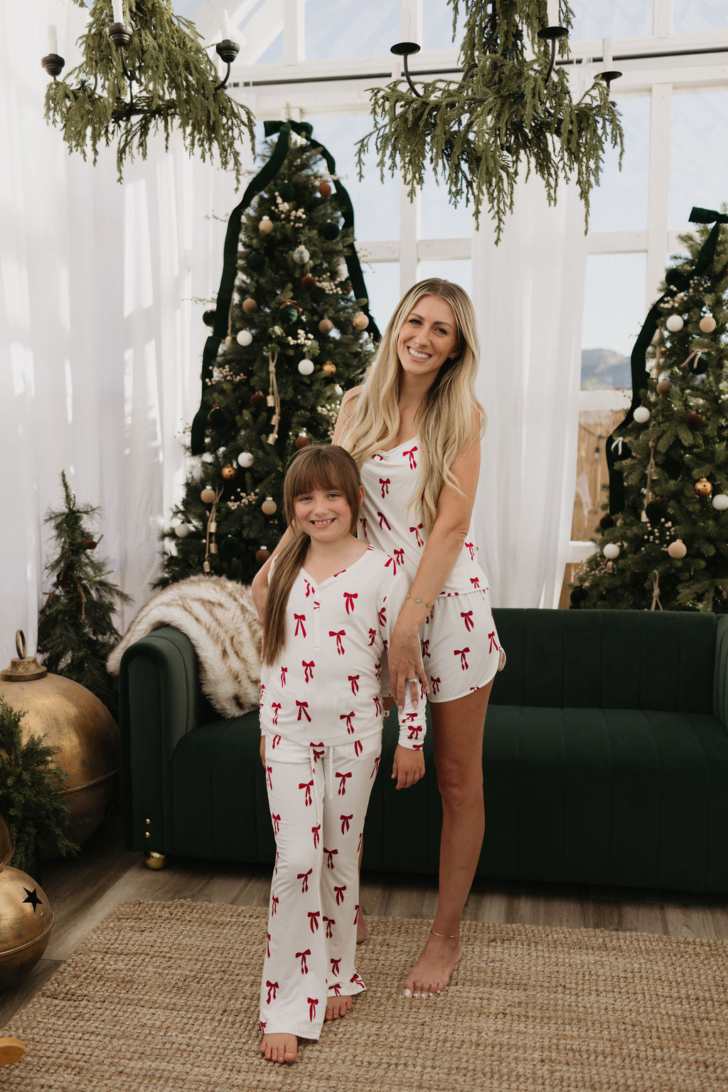 A woman and a young girl are smiling and wearing matching pajamas, including a set of "Pre-Teen Flare Bamboo Pajamas" in the delightful "Ribbons & Bows" pattern by forever french baby. They stand in a festive room adorned with Christmas trees, ornaments, and greenery, ideal for enjoying hypo-allergenic clothing. A dark green couch is visible in the background.