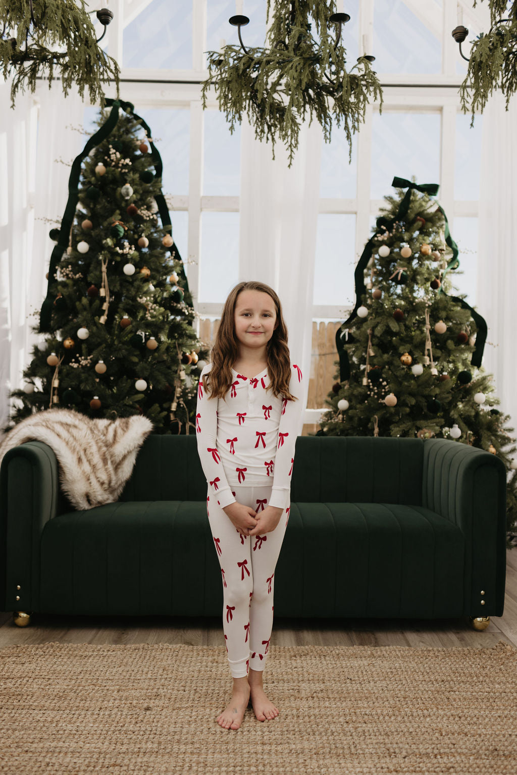 A child wearing the "Bamboo Two Piece Pajamas | Ribbons & Bows" by forever french baby stands barefoot in front of a green sofa. Behind them, two decorated Christmas trees and ceiling-hung greenery contribute to a cozy and festive atmosphere.