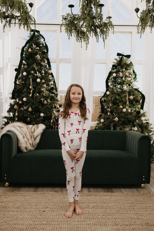 A child wearing the "Bamboo Two Piece Pajamas | Ribbons & Bows" by forever french baby stands barefoot in front of a green sofa. Behind them, two decorated Christmas trees and ceiling-hung greenery contribute to a cozy and festive atmosphere.