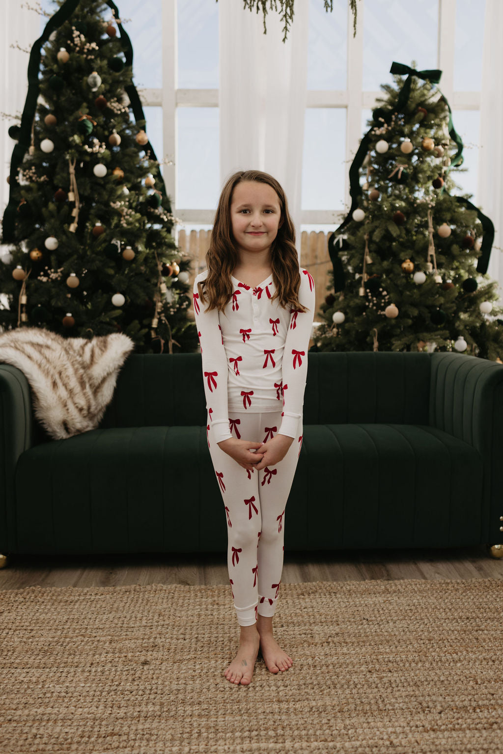 A young girl in forever french baby hypo-allergenic Bamboo Two Piece Pajamas, called Ribbons & Bows, stands barefoot on a woven rug. She is in front of a dark green sofa, with two decorated Christmas trees in the background by a large window. Her pajamas, accented with red bows, add to the festive scene.