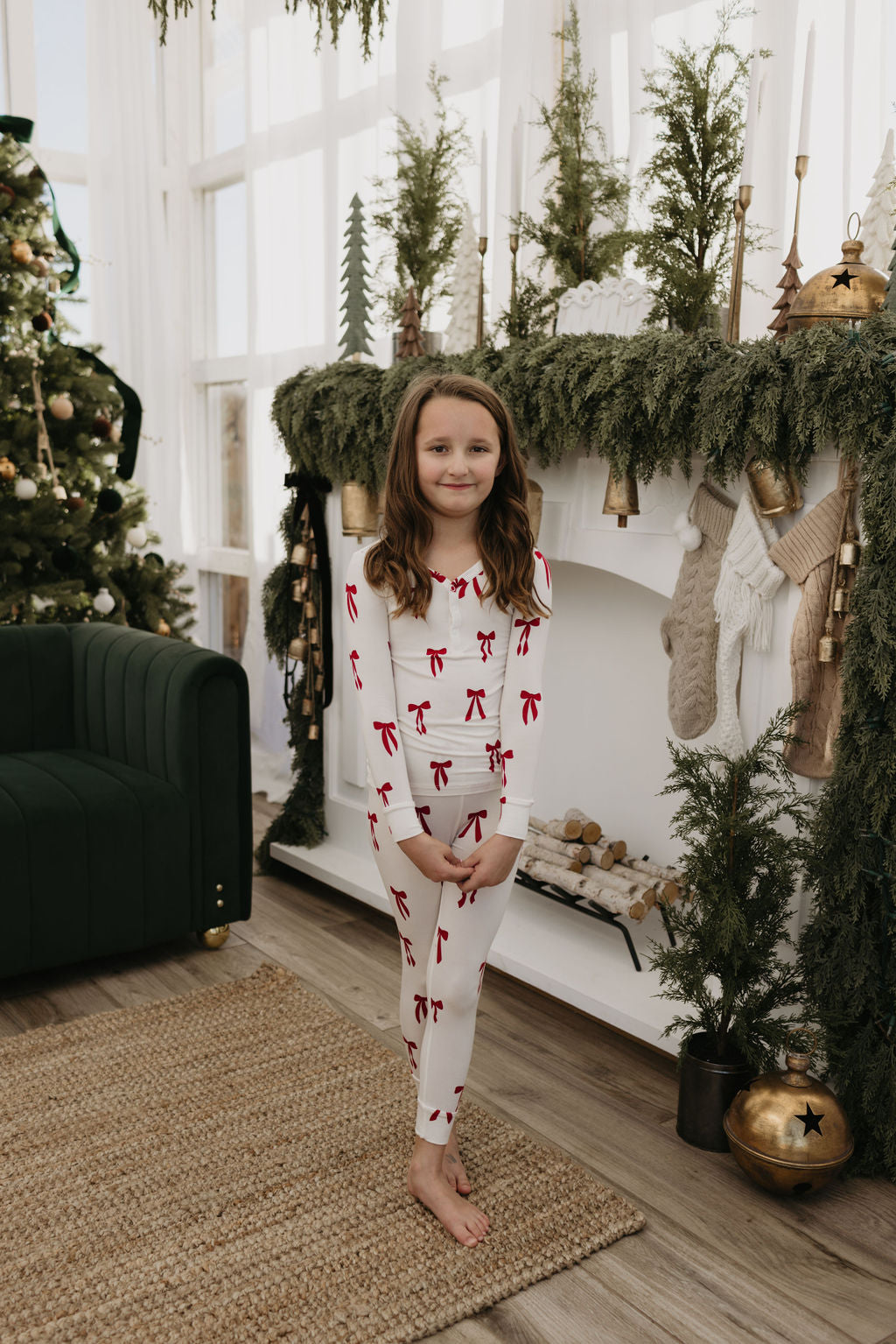 A young girl wearing the Bamboo Two Piece Pajamas in the Ribbons & Bows design from forever french baby stands barefoot and smiling before a decorated fireplace with stockings and greenery. Her festive, hypo-allergenic sleepwear enhances the scene, while an ornament-adorned Christmas tree is visible in the background.