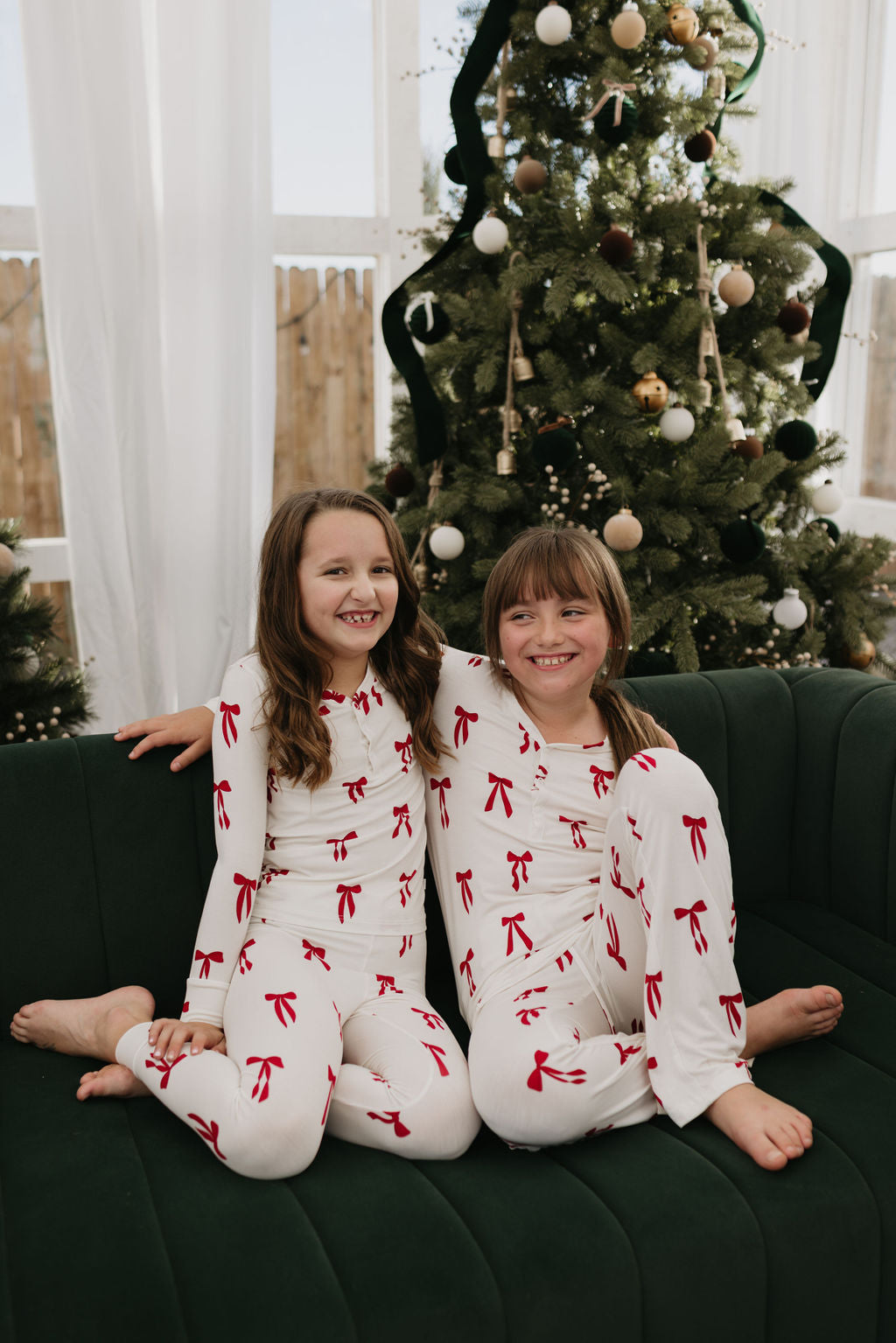 Two pre-teens in matching "Pre-Teen Flare Bamboo Pajamas" by forever french baby, featuring the Ribbons & Bows design in white with red bows, sit smiling on a green couch. A decorated Christmas tree stands nearby, creating a festive atmosphere with ornaments and ribbons.