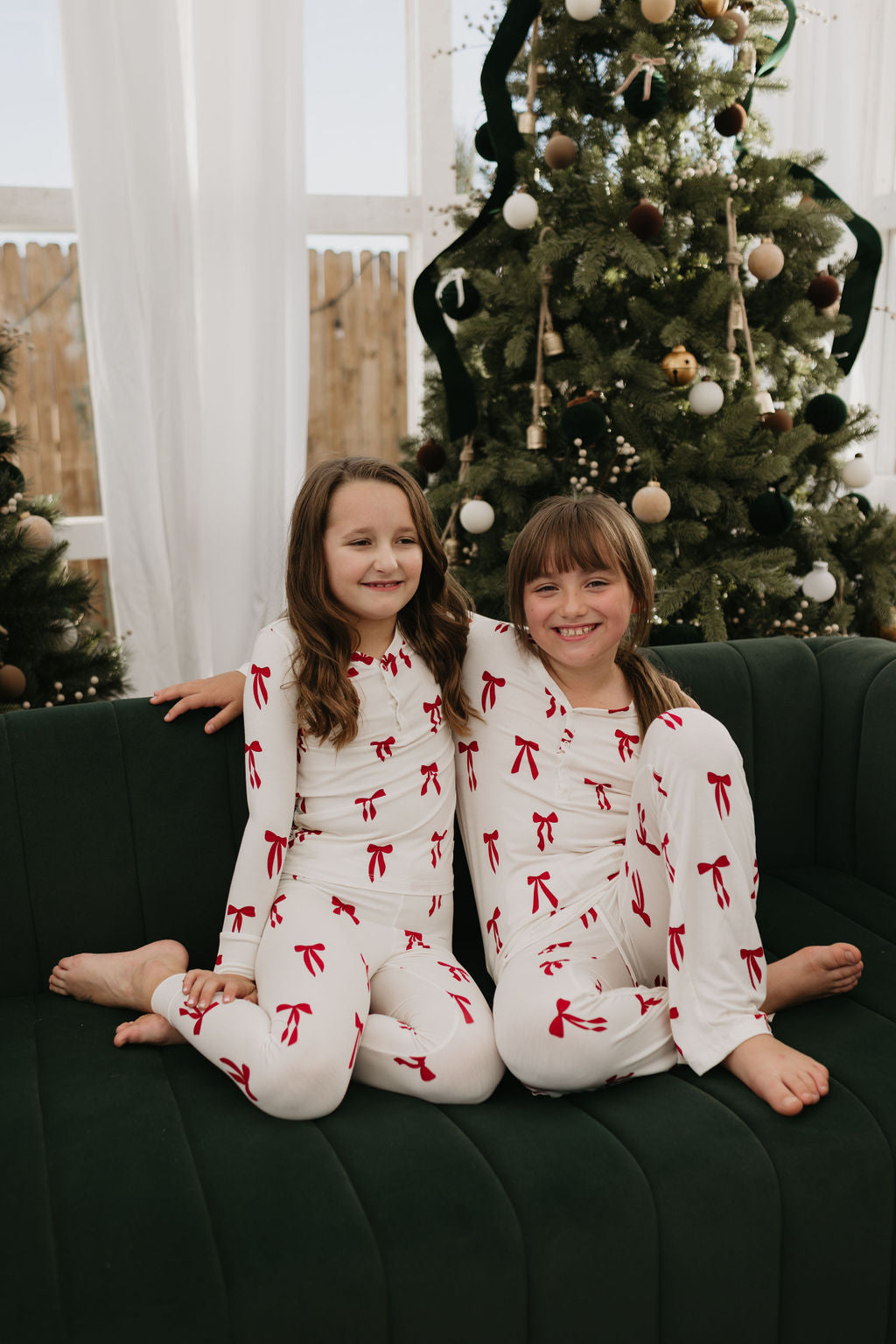 Two children sit on a dark green sofa wearing matching forever french baby Pre-Teen Flare Bamboo Pajamas in the Ribbons & Bows pattern. They smile happily, with a Christmas tree decorated in the background.