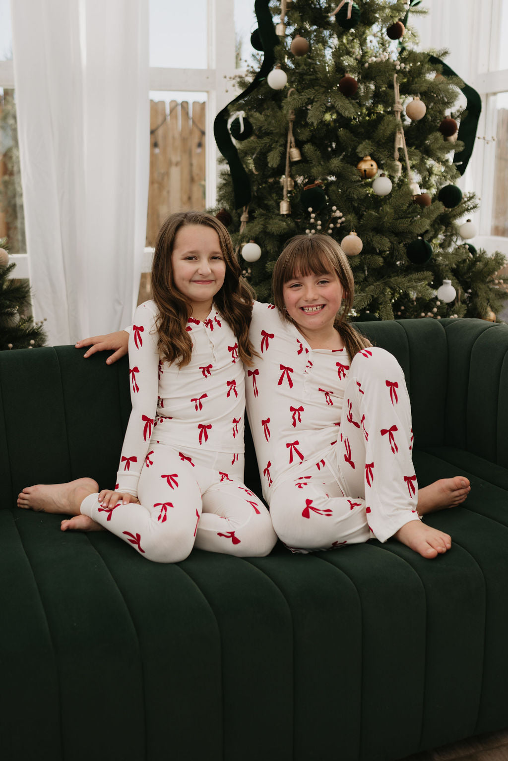 Two children wearing forever french baby's Bamboo Two Piece Pajamas in the Ribbons & Bows design are seated on a green sofa in front of a decorated Christmas tree. They are smiling and seem to be enjoying a festive moment indoors.