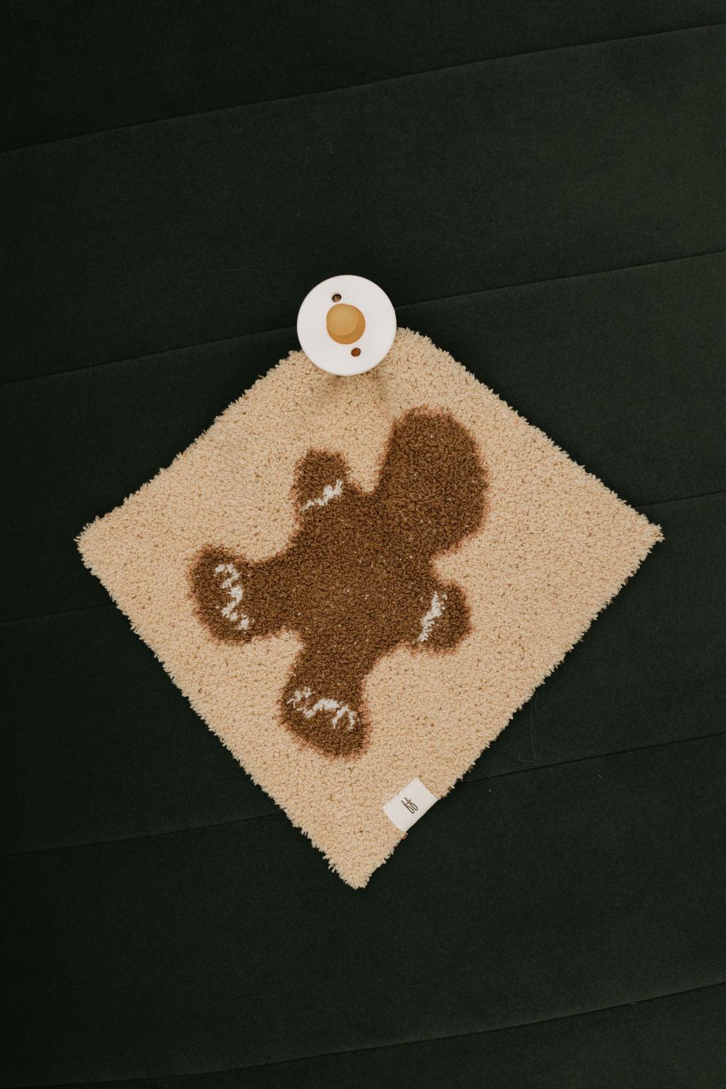 As part of forever french baby's Gingerbread Collection, the Lovey teether features a plush, diamond-shaped beige design adorned with a brown gingerbread man motif and rests gracefully on a dark green quilted background. A small round cup with a beige lid sits neatly at the top corner of this delightful piece.