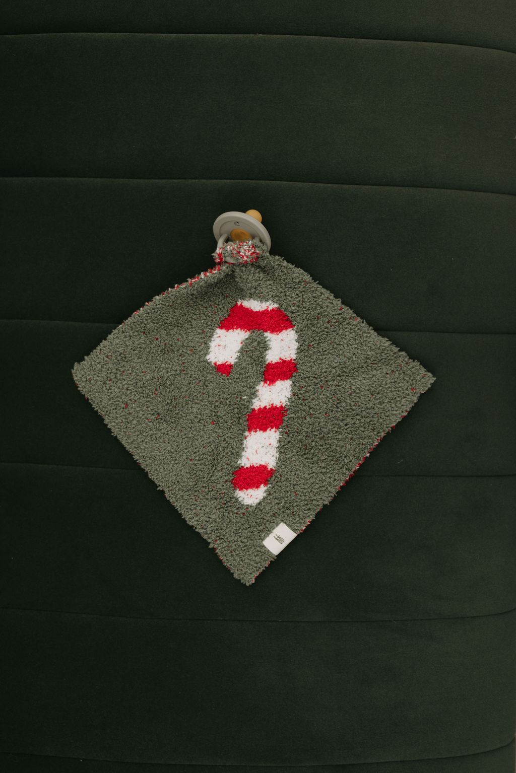 The Lovey | Candy Cane Lane by Forever French Baby is a luxurious, gray square cloth made from microfiber feather yarn. It showcases a red and white striped candy cane design attached to a pacifier, all set against a dark green background with vertical stitching. This elegant accessory is ideal for any Forever French Baby collection.