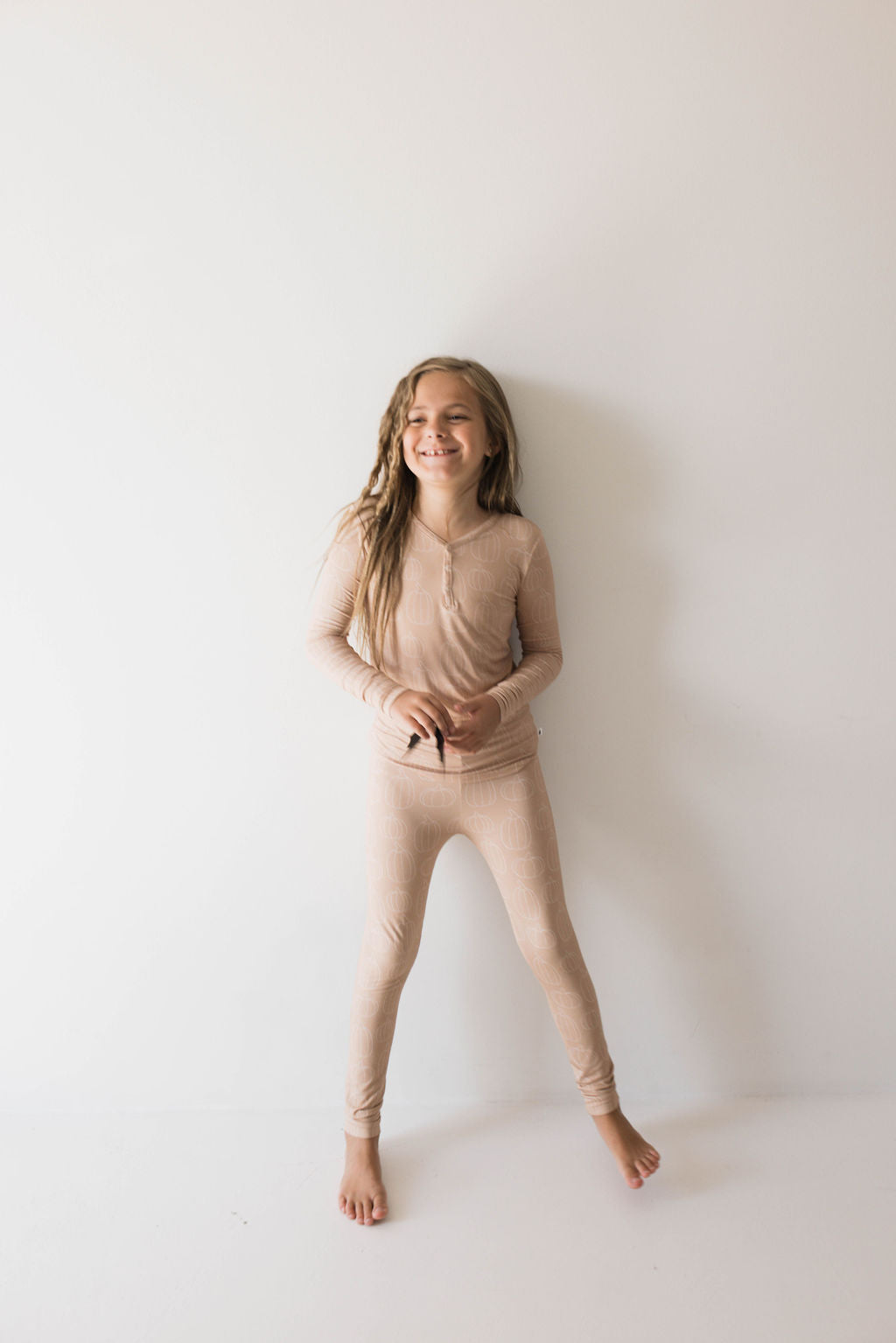 A young girl is standing barefoot against a plain light-colored wall. She is smiling and wearing matching light beige Bamboo Two Piece Pajamas in the Pumpkin Pie design from forever french baby, resembling thermal or loungewear. Her hair is long and slightly wavy, and she appears relaxed and happy, showcasing the comfort of hypo-allergenic children's sleepwear.