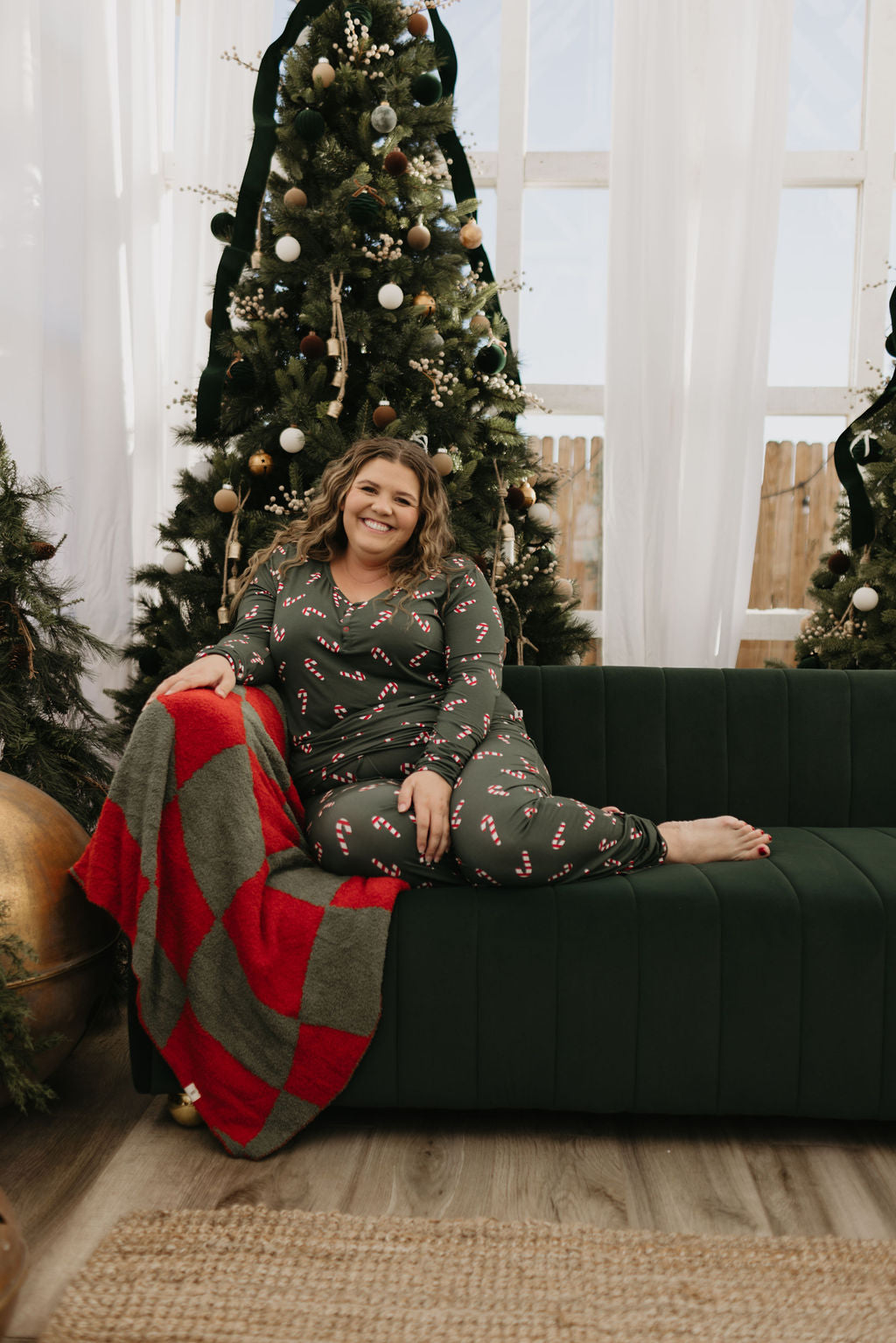 A person wearing festive Women's Bamboo Pajamas from forever french baby, designed with candy cane patterns and hypo-allergenic properties, sits on a dark green sofa. They smile contentedly with beautifully decorated Christmas trees in the background. A red and green blanket is draped over the sofa, offering ideal comfort for breastfeeding moms during the holidays.