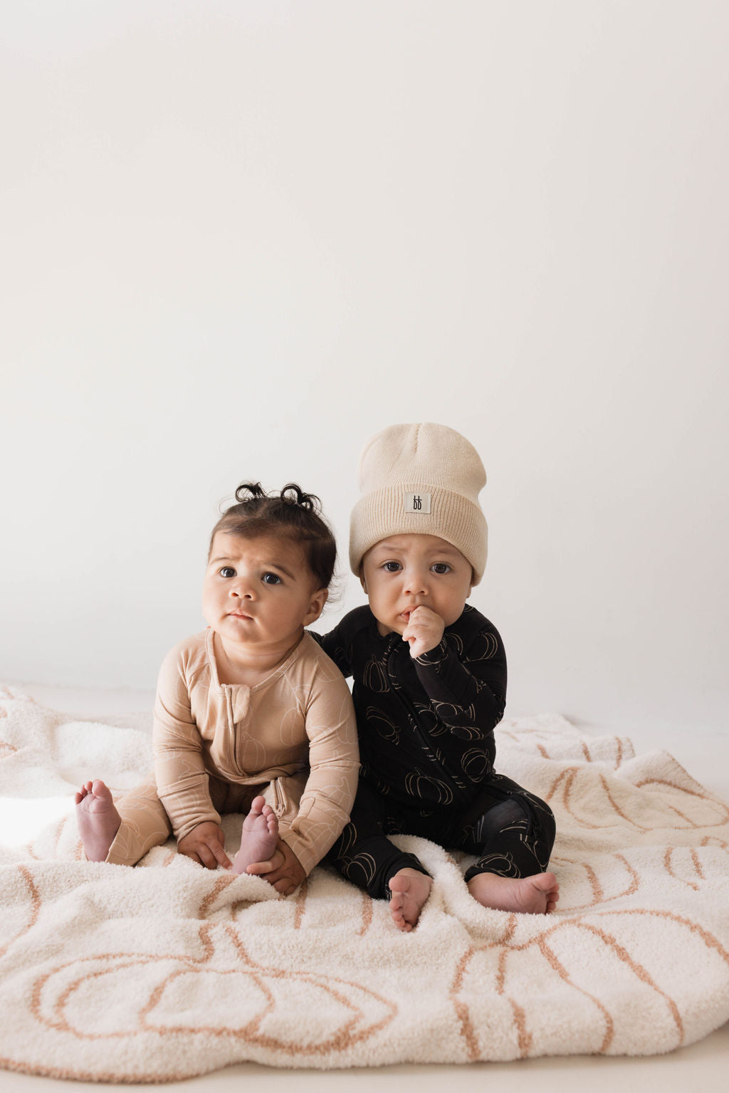 Two babies sit on a patterned blanket against a plain background. The baby on the left wears "Pumpkin Pie" Bamboo Zip Pajamas by forever french baby and looks slightly off-camera. The baby on the right wears a black outfit with a light-colored beanie, holding one hand to their mouth.