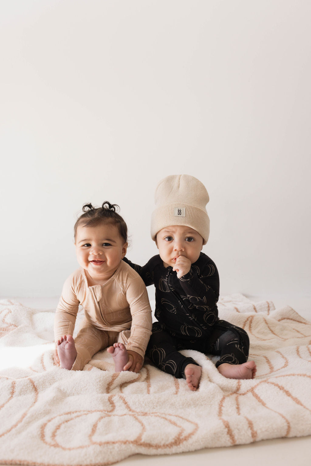 Two babies are seated on a soft, patterned blanket. The baby on the left wears the Bamboo Zip Pajamas in Pumpkin Pie by forever french baby and smiles at the camera, while the baby on the right wears a black onesie with gold designs and a beige beanie, looking thoughtful with a finger near their mouth.