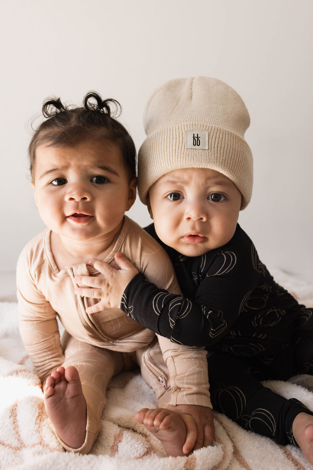 Two babies sit close together, both looking towards the camera. The baby on the left wears a light-colored, breathable outfit with curly hair styled into two little pigtails. The baby on the right sports forever french baby's Bamboo Zip Pajamas in Pumpkin Pie along with a beige beanie and has their arm around the other baby.