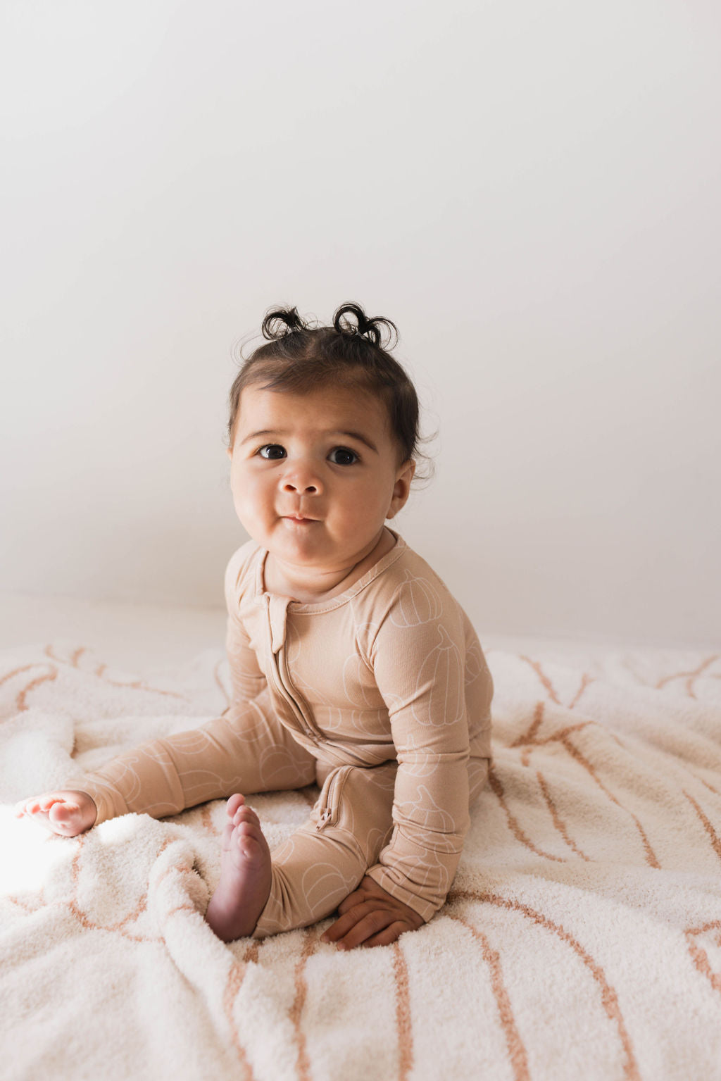 A baby with dark hair sits on a soft, patterned blanket. The baby is wearing forever french baby's Bamboo Zip Pajamas in the Pumpkin Pie design, characterized by its hypo-allergenic beige fabric with a subtle pattern. The baby looks ahead with a calm expression against a neutral, light-colored background.