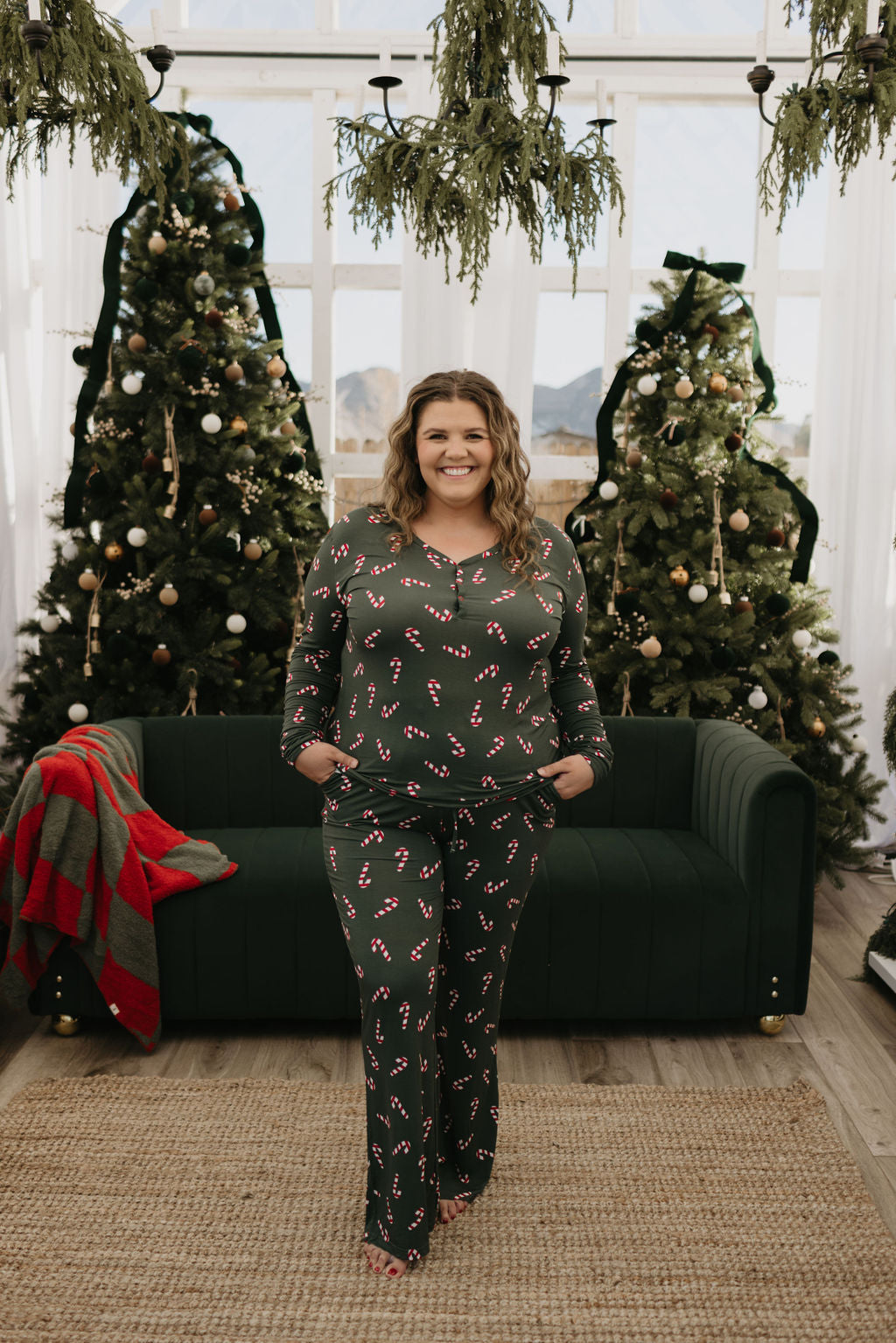 A person wearing the Women's Bamboo Pajamas from forever french baby's Candy Cane Lane collection stands in a festive room with decorated Christmas trees. A green sofa with a red and green blanket offers comfort, ideal for breastfeeding moms. Hanging greenery adorns the ceiling, enhancing the cozy holiday vibe.