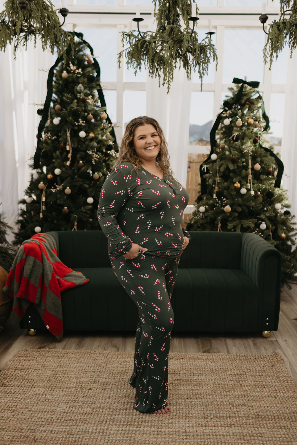 Standing in front of a dark green sofa, a person wears the Women's Bamboo Pajamas in Candy Cane Lane from forever french baby. Behind them, three decorated Christmas trees with white ornaments fill the room, which features large windows and hanging greenery—an ideal setting for cozy moments for breastfeeding moms.