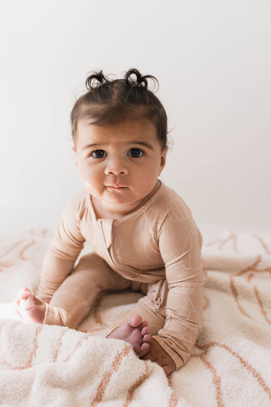 A baby with dark curly hair sits on a soft, patterned blanket. The baby wears the Bamboo Zip Pajamas in Pumpkin Pie by forever french baby, made of breathable fabric and looks directly at the camera with a curious expression. The background is softly lit, creating a warm and cozy atmosphere.